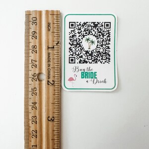 May include: A white square sticker with a black and white QR code, a pink flamingo, and green palm trees. The text "Buy the BRIDE a Drink" is written in green cursive.