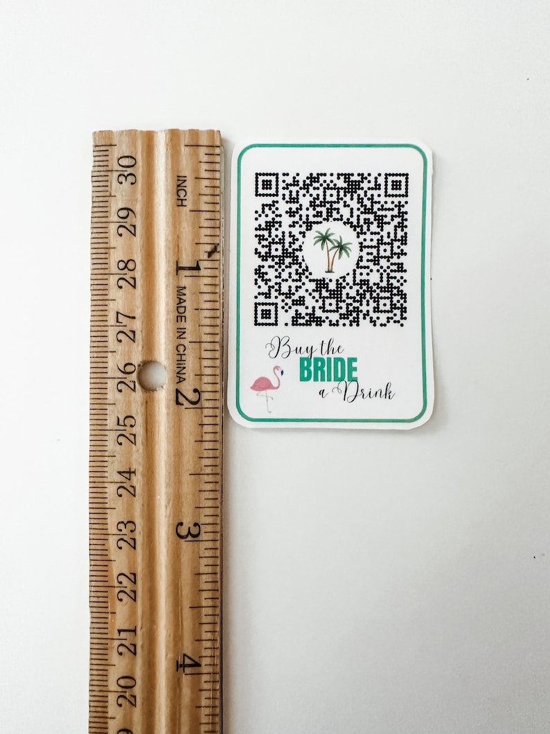 May include: A white square sticker with a black and white QR code, a pink flamingo, and green palm trees. The text "Buy the BRIDE a Drink" is written in green cursive.