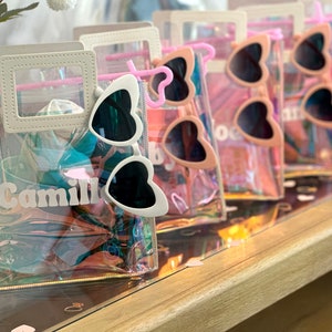 May include: Clear iridescent plastic tote bags with a white handle and a white vinyl name tag. The bags contain heart-shaped sunglasses with black lenses. One bag has the name "Camille" on the name tag.