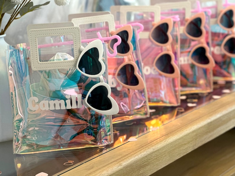 May include: Clear iridescent plastic tote bags with a white handle and a white vinyl name tag. The bags contain heart-shaped sunglasses with black lenses. One bag has the name "Camille" on the name tag.
