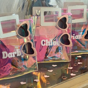 May include: Four clear plastic bags with pink heart-shaped sunglasses inside. Each bag has a white top flap with a name written on it: Darby, Chloe, Arian. The bags are on a wooden surface with pink heart-shaped confetti.