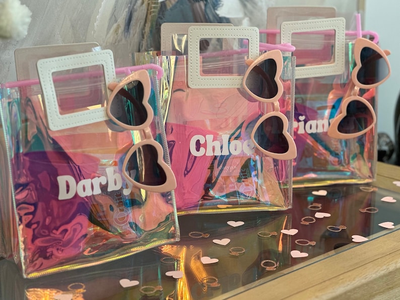 May include: Four clear plastic bags with pink heart-shaped sunglasses inside. Each bag has a white top flap with a name written on it: Darby, Chloe, Arian. The bags are on a wooden surface with pink heart-shaped confetti.