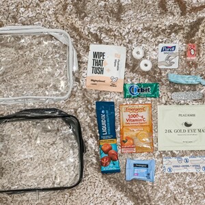 May include: Two clear plastic toiletry bags alongside various travel-sized items including a Wipe That Tush packet, two mints, a Purell wipe, a small red item, a blue hair tie, a nail file, Orbit gum, Liquid I.V. stick, Emergen-C packet, Peauamie eye mask, Neutrogena wipe, and a sterile bandage.