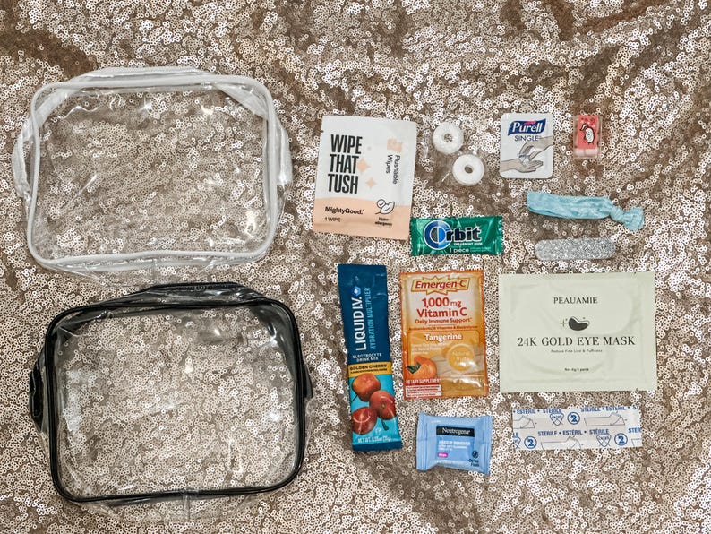 May include: Two clear plastic toiletry bags alongside various travel-sized items including a Wipe That Tush packet, two mints, a Purell wipe, a small red item, a blue hair tie, a nail file, Orbit gum, Liquid I.V. stick, Emergen-C packet, Peauamie eye mask, Neutrogena wipe, and a sterile bandage.
