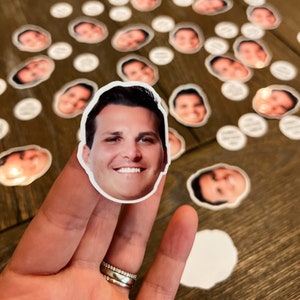 Pu&ograve; includere: Un primo piano di un adesivo bianco con una foto del viso di una persona. L'adesivo &egrave; tenuto da una mano con un anello al dito. L'adesivo &egrave; su una superficie di legno con altri adesivi sparsi intorno.