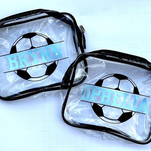 Peut inclure: Deux trousses rectangulaires transparentes avec des bordures et des fermetures éclair noires. Chaque trousse présente un motif de ballon de football noir et blanc avec une bande horizontale. Le nom BRYNN est imprimé sur l'une et OPHELIA sur l'autre.