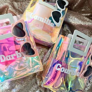 May include: Three iridescent clear plastic bags with white handles and white lettering. The bags are filled with various items, including sunglasses, a pink hairbrush, and a pink bottle. The bags are labelled "Camille", "Darby", and "Cournte".