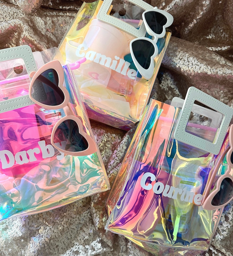 May include: Three iridescent clear plastic bags with white handles and white lettering. The bags are filled with various items, including sunglasses, a pink hairbrush, and a pink bottle. The bags are labelled "Camille", "Darby", and "Cournte".