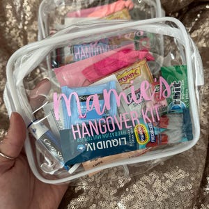 May include: A clear rectangular vinyl pouch with white trim containing various small personal care items, including gum, lip balm, and hydration packets. The front of the pouch features the pink text "mamie's HANGOVER KIT" printed on the surface.