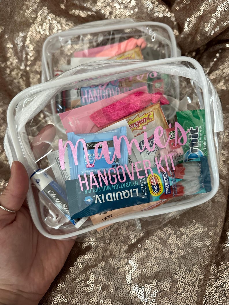 May include: A clear rectangular vinyl pouch with white trim containing various small personal care items, including gum, lip balm, and hydration packets. The front of the pouch features the pink text "mamie's HANGOVER KIT" printed on the surface.