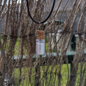 May include: A silver necklace with a small glass bottle filled with iridescent glitter. The bottle has a cork stopper and is hanging from a black cord.