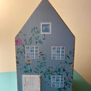May include: A decorative wooden house painted in a light blue hue, adorned with floral vines and roses. The house features three white-framed windows, a small door, and a single window at the top. The design is reminiscent of a cottage or farmhouse.