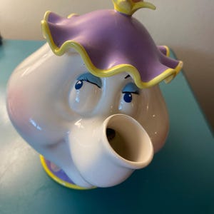 May include: A white ceramic teapot with a cartoon face, featuring a purple lid with yellow trim and a pink sphere on top. The teapot has blue eyes and a spout. The base has a yellow and purple design.