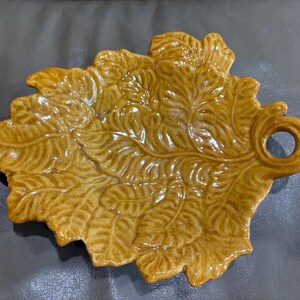 May include: A glossy, mustard-yellow ceramic dish shaped like a large, textured leaf. The surface features intricate, raised vein patterns throughout the foliage design, and the stem end forms a small, circular handle loop on the right side.