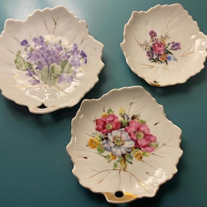 Puede incluir: Tres platos blancos de porcelana con forma de hoja y ribete dorado. Cada plato presenta un diseño floral diferente en tonos morados, rosas y amarillos. Los platos tienen una pequeña abertura de asa en la base.