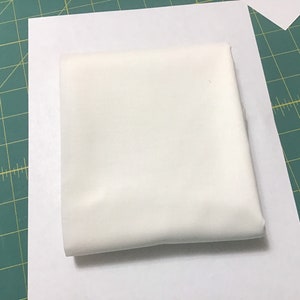 May include: A folded, square piece of white fabric, possibly cotton or linen, rests on a white sheet of paper. The fabric appears smooth and opaque, with soft shadows suggesting its folded form. The background is a green cutting mat.