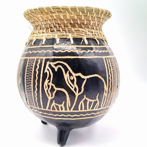 May include: A black ceramic pot with three legs and a woven straw lid. The pot is decorated with a carved design of elephants.