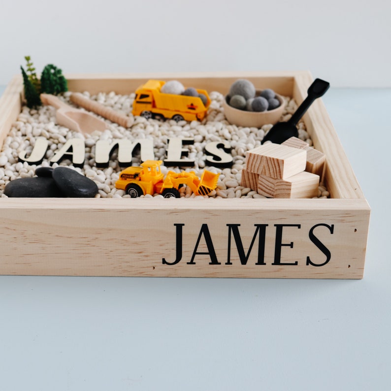 May include: A wooden sensory bin with the name "JAMES" spelled out in black letters. The bin is filled with white beans, small grey rocks, and toy construction vehicles.
