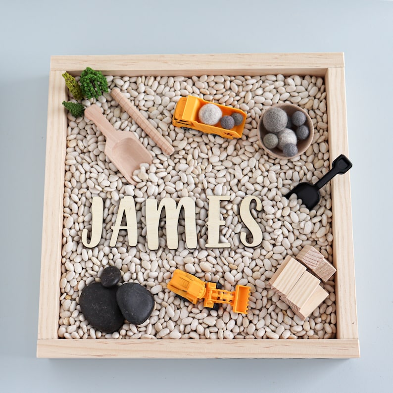 May include: A wooden tray filled with white beans, featuring a name spelled out in wooden letters, "JAMES".  The tray also includes toy construction vehicles, a small shovel, and a pile of wooden blocks.