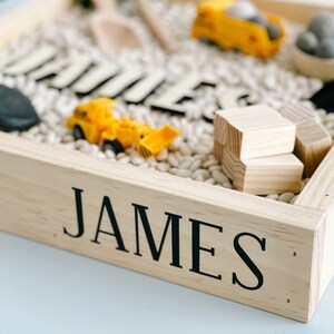 May include: A wooden sensory bin with the name "JAMES" on the front. The bin is filled with white beans and includes toy construction vehicles, wooden blocks, and letter tiles.