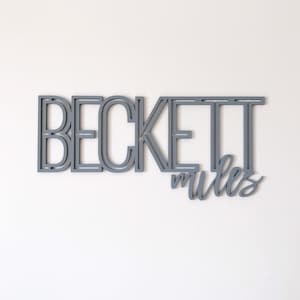 Puede incluir: Un letrero de madera gris con el nombre "Beckett Miles" en una fuente en capas y dimensional.