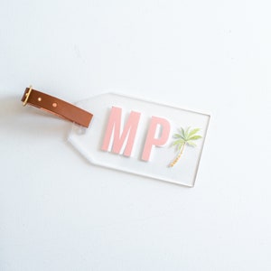 May include: Clear luggage tag with a brown leather strap. The tag has the letters "MP" in pink and a small watercolor palm tree.