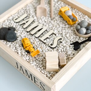 May include: A wooden sensory bin with the name "JAMES" spelled out in black letters. The bin is filled with white beans and includes toy construction vehicles, wooden blocks, and a small shovel.