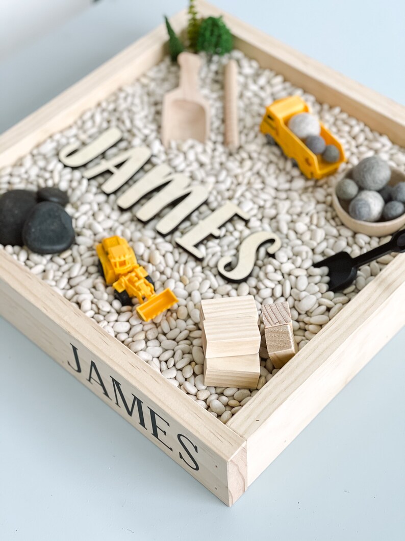 May include: A wooden sensory bin with the name "JAMES" spelled out in black letters. The bin is filled with white beans and includes toy construction vehicles, wooden blocks, and a small shovel.
