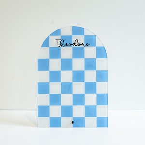 May include: A blue and white checkered arch-shaped sign with the name "Theodore" written in black.