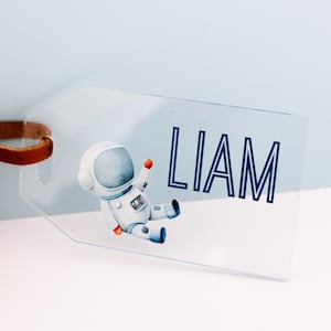 May include: A clear plastic luggage tag with a blue and white astronaut illustration and the name "LIAM" printed in blue.