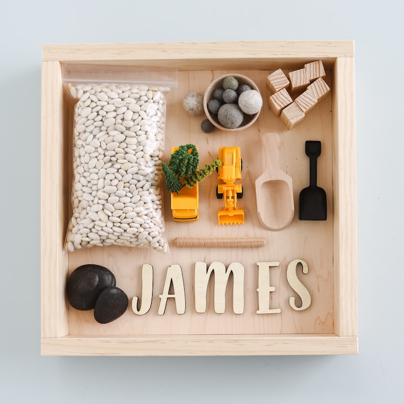 May include: A wooden box with a light wood finish contains a variety of items for a sensory bin. The items include white beans, small wooden blocks, a small shovel, a small scoop, a toy truck, a small plant, and two gray stones. The name "JAMES" is spelled out in large wooden letters.