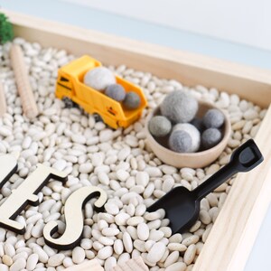 May include: A wooden sensory bin filled with white beans, a yellow toy dump truck, and a black plastic shovel. The bin also contains a small bowl filled with gray felt balls.