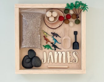 May include: A wooden sensory bin with the name "JAMES" on the front. The bin contains a variety of items, including rice, dinosaurs, rocks, a wooden scoop, a small shovel, and a palm tree.