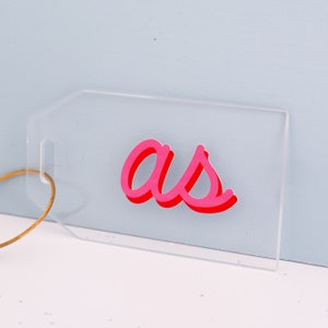 May include: A clear acrylic luggage tag with the word "as" written in pink and red lettering.
