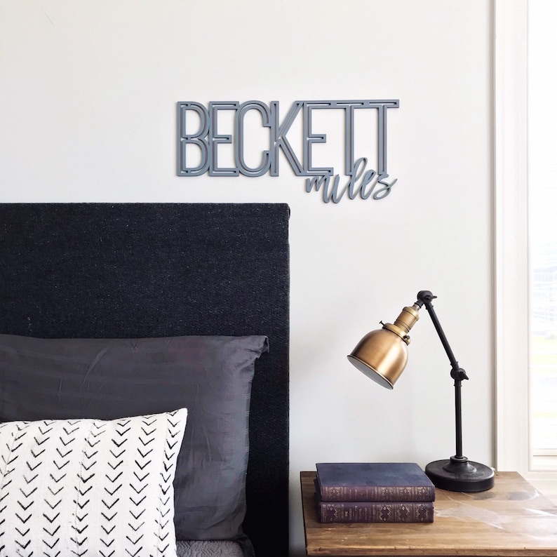 Puede incluir: Un letrero de madera gris con el nombre "Beckett" y las palabras "rules" escritas en cursiva. El letrero est&aacute; colgado en una pared blanca sobre una cabecera negra.