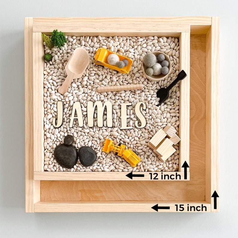 May include: A wooden sensory bin with a white bean filler. The bin is 15 inches wide and 12 inches deep. The bin includes a variety of small toys, including a toy truck, a shovel, and rocks. The name "JAMES" is spelled out in wooden letters.
