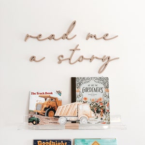 May include: Wooden wall decor with the words "read me a story" in cursive script.  The decor is displayed above a shelf with three children's books: "The Truck Book", "We Are The Gardeners", and "Goodnight".