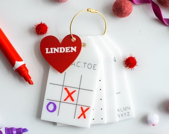 May include: A white card with a red heart that says "Linden" on it. The card is attached to a gold chain and has a tic-tac-toe game on it. The game is partially played with an 'O' and two 'X's. The card also has a section with the letters "KLMN" and "XYZ" on it.