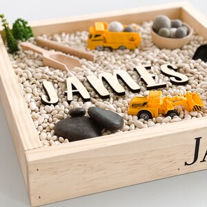 May include: A wooden sensory bin with white beans, black stones, and toy construction vehicles. The name "JAMES" is spelled out in wooden letters.