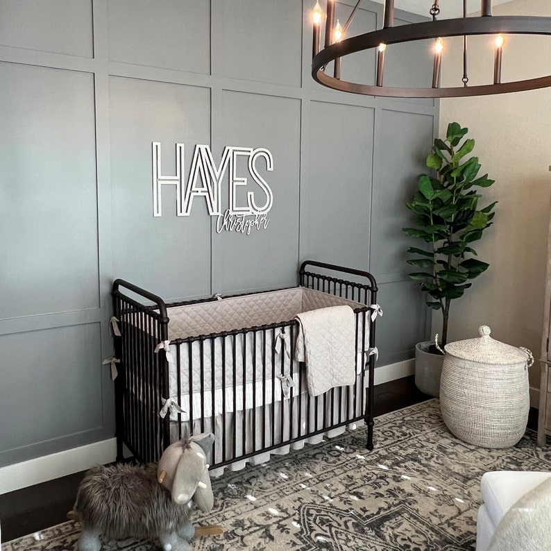 Puede incluir: Una habitaci&oacute;n de beb&eacute; con una cuna de metal negro, una alfombra gris y blanca, una cesta de mimbre blanca y un animal de peluche gris y blanco. La pared est&aacute; pintada de gris y tiene un letrero de madera blanco con el nombre "HAYES Christopher".