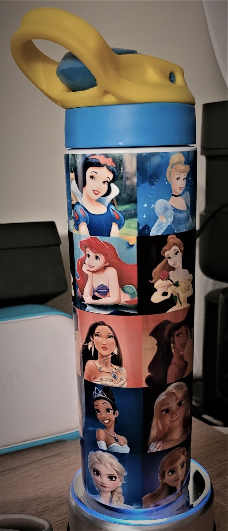 May include: A tall water bottle featuring images of Disney princesses. The bottle has a blue base and a yellow handle. The princesses include Snow White, Cinderella, Ariel, Belle, Pocahontas, Tiana, and Elsa.