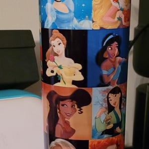 May include: A tall, insulated water bottle featuring images of Disney princesses. The bottle has a blue and yellow lid and a white body with images of Cinderella, Aurora, Belle, Jasmine, and others. The bottle is designed to keep drinks cold.