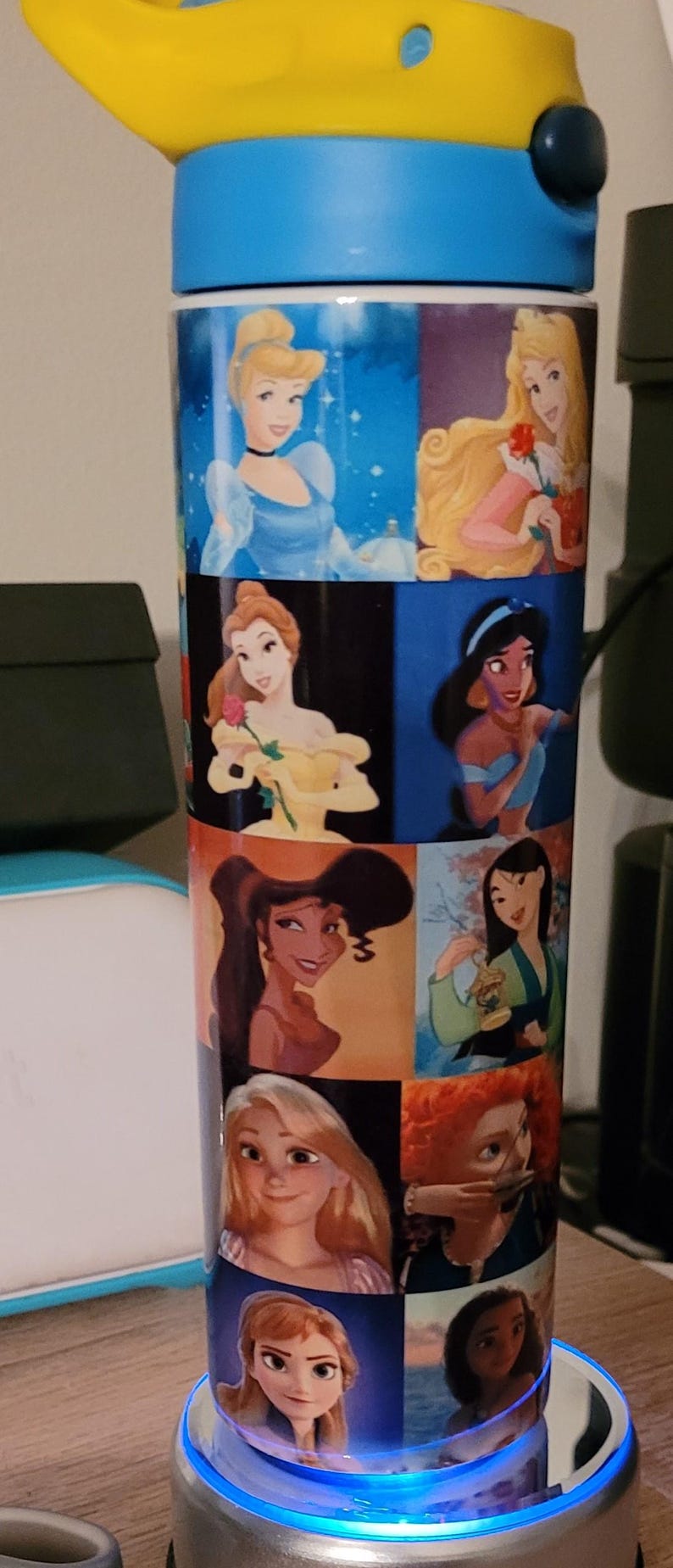 May include: A tall, insulated water bottle featuring images of Disney princesses. The bottle has a blue and yellow lid and a white body with images of Cinderella, Aurora, Belle, Jasmine, and others. The bottle is designed to keep drinks cold.