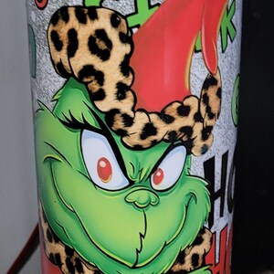 May include: A green Grinch character with red eyes and a leopard print hat is featured on a white mug with the words "Stink Stunk" in red and black lettering. The mug is on a silver base with a light blue and purple iridescent ring around the bottom.