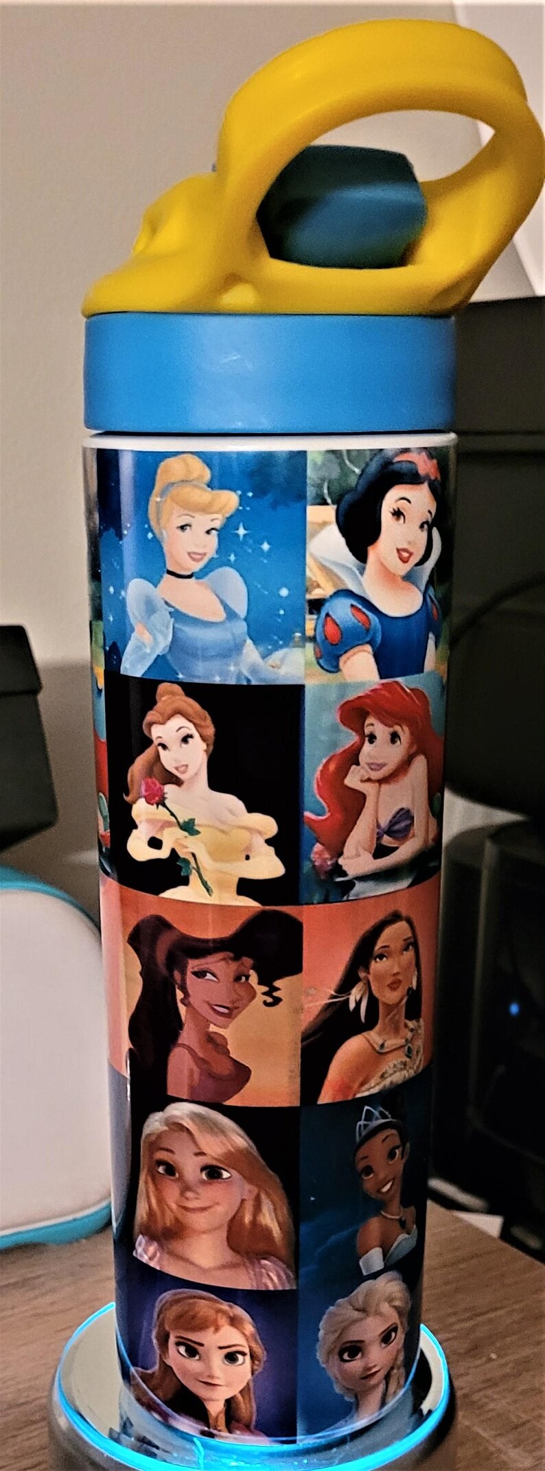 May include: A tall, cylindrical water bottle featuring images of Disney princesses. The bottle has a blue base and lid, with a yellow handle. The princesses include Cinderella, Snow White, Belle, Ariel, and others. The bottle is designed for hydration.