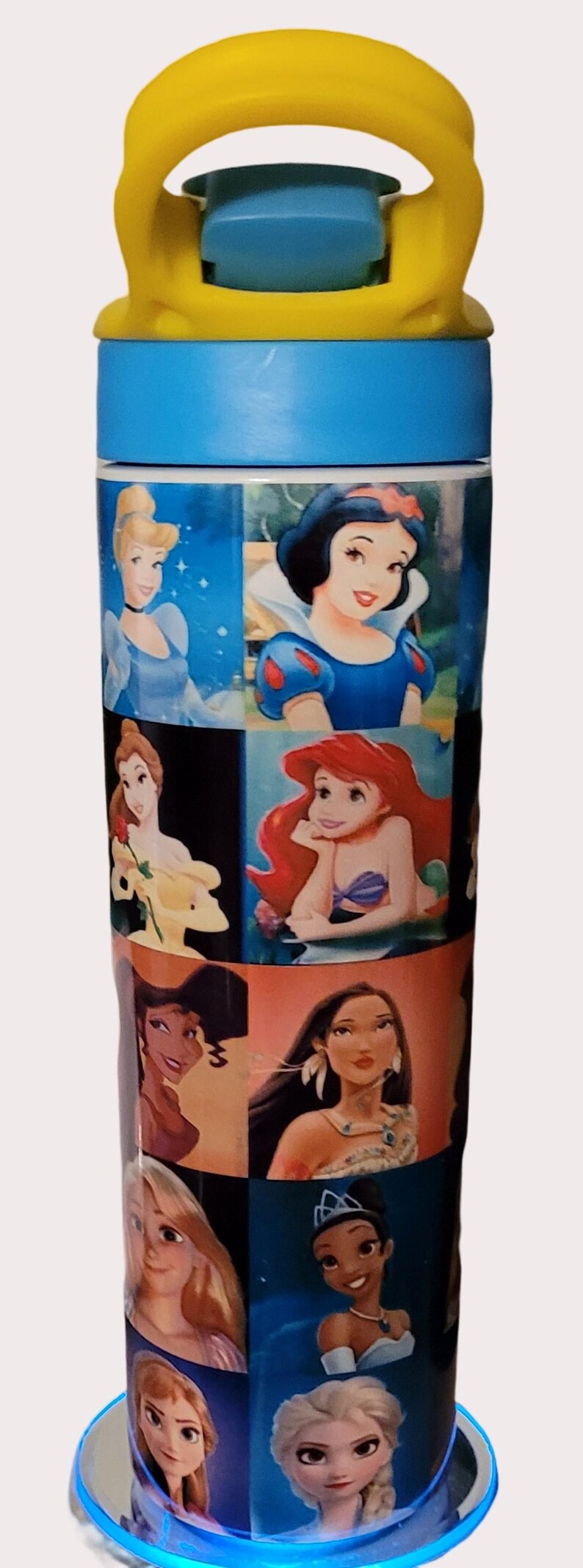 May include: A tall, cylindrical water bottle featuring images of Disney princesses. The bottle has a blue top and base, a yellow handle, and a series of princess portraits, including Cinderella, Snow White, and Ariel.