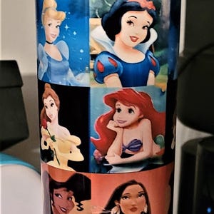 May include: A blue and yellow water bottle featuring images of Disney princesses. The bottle has a yellow handle and a blue lid. The images include Cinderella, Snow White, Ariel, and others. The bottle is cylindrical.