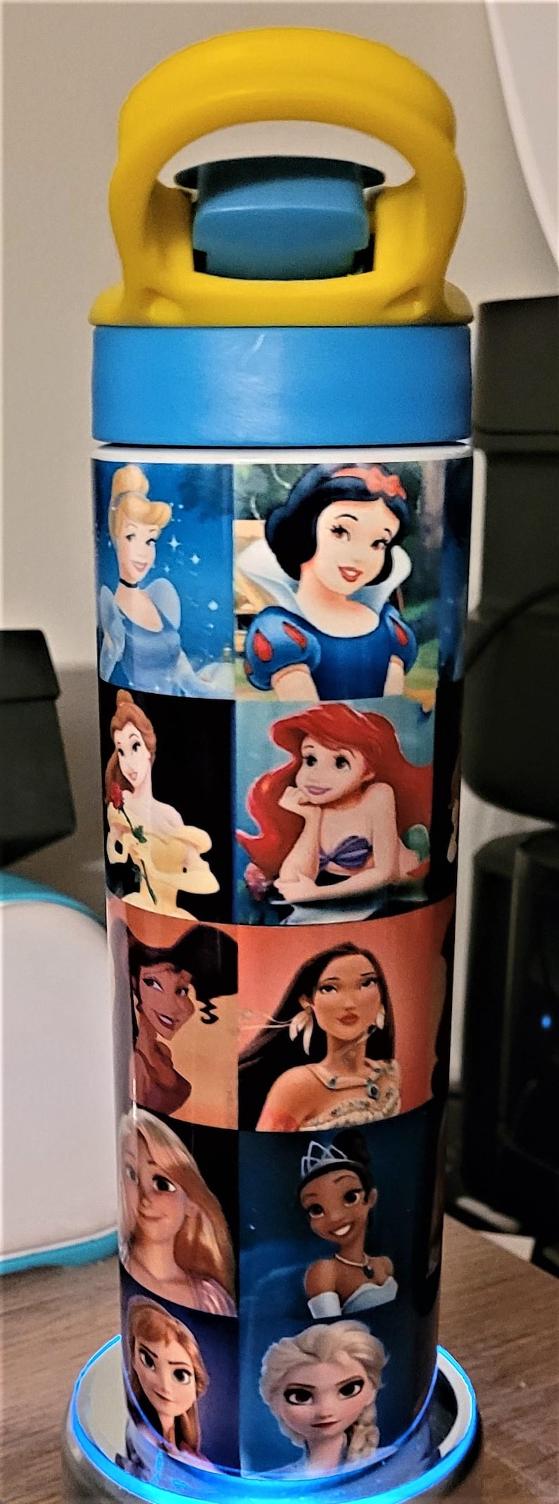 May include: A blue and yellow water bottle featuring images of Disney princesses. The bottle has a yellow handle and a blue lid. The images include Cinderella, Snow White, Ariel, and others. The bottle is cylindrical.