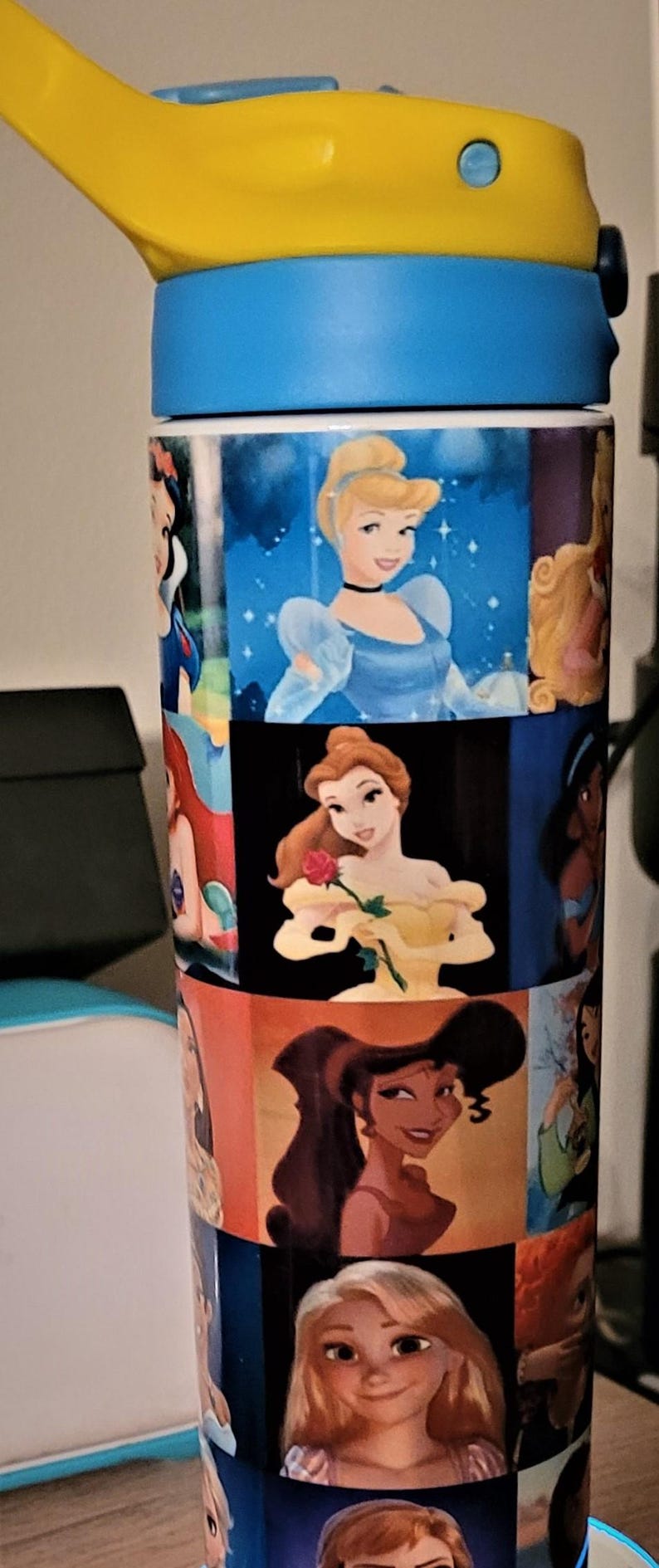 May include: A blue and yellow water bottle featuring images of Disney princesses. The bottle has a yellow flip-top lid and a blue base. The body of the bottle is covered in colorful images of Cinderella, Belle, and other Disney princesses.