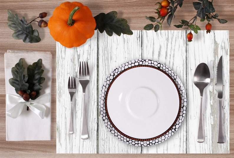 Puede incluir: Un mantel individual blanco de imitaci&oacute;n de madera con un plato blanco, un tenedor, una cuchara y un cuchillo. El mantel individual tiene un aspecto desgastado con pintura blanca sobre un patr&oacute;n de veta de madera. Hay una peque&ntilde;a calabaza y una rama de hojas en el fondo.